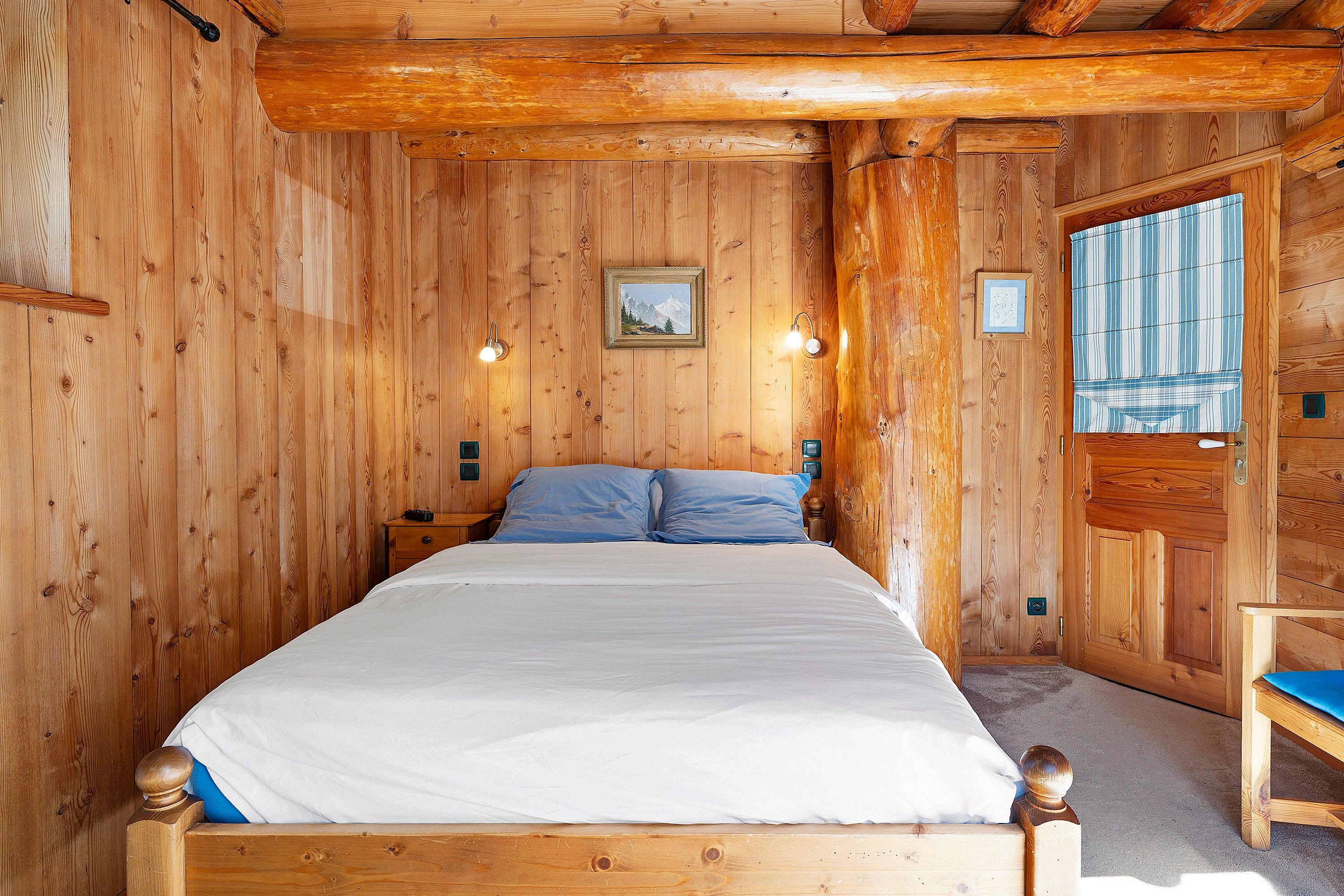 A wooden room with a bed, a nightstand, a wall-mounted lamp, and a door with a window.