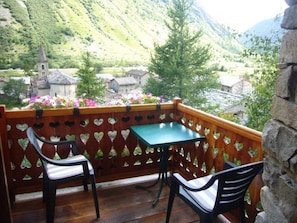 A balcony with a table and two chairs, overlooking a village with a church and mountains. - Le Marmotton Gîte – Chalet Les Abeillos, Mountain View Retreat for 2 (Bonneval-sur-Arc)