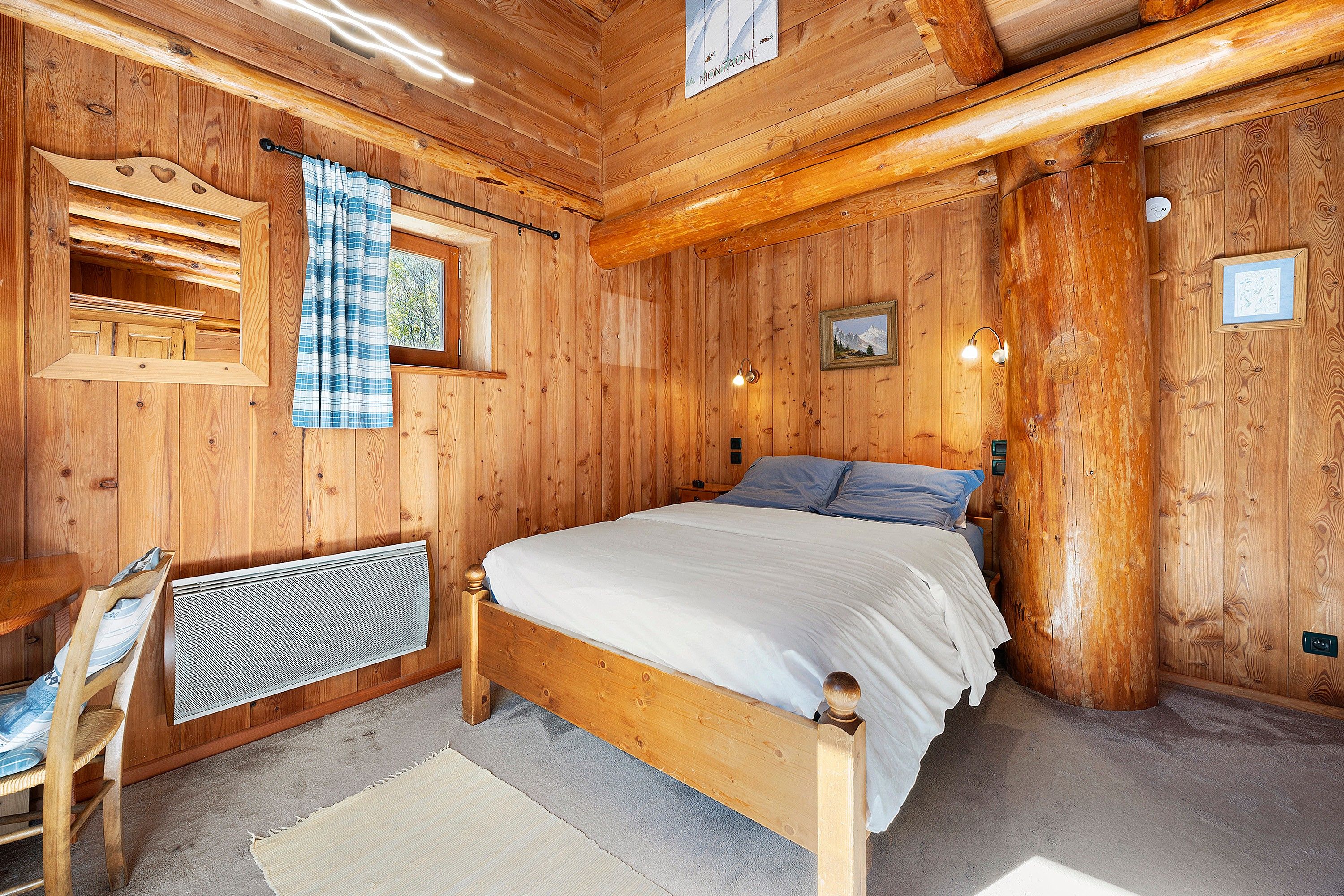 Une chambre dans une cabane en bois, avec un lit, une fenêtre avec des rideaux, un tableau accroché au mur et un radiateur.