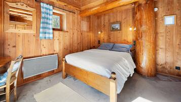 Une chambre dans une cabane en bois, avec un lit, une fenêtre avec des rideaux, un tableau accroché au mur et un radiateur.