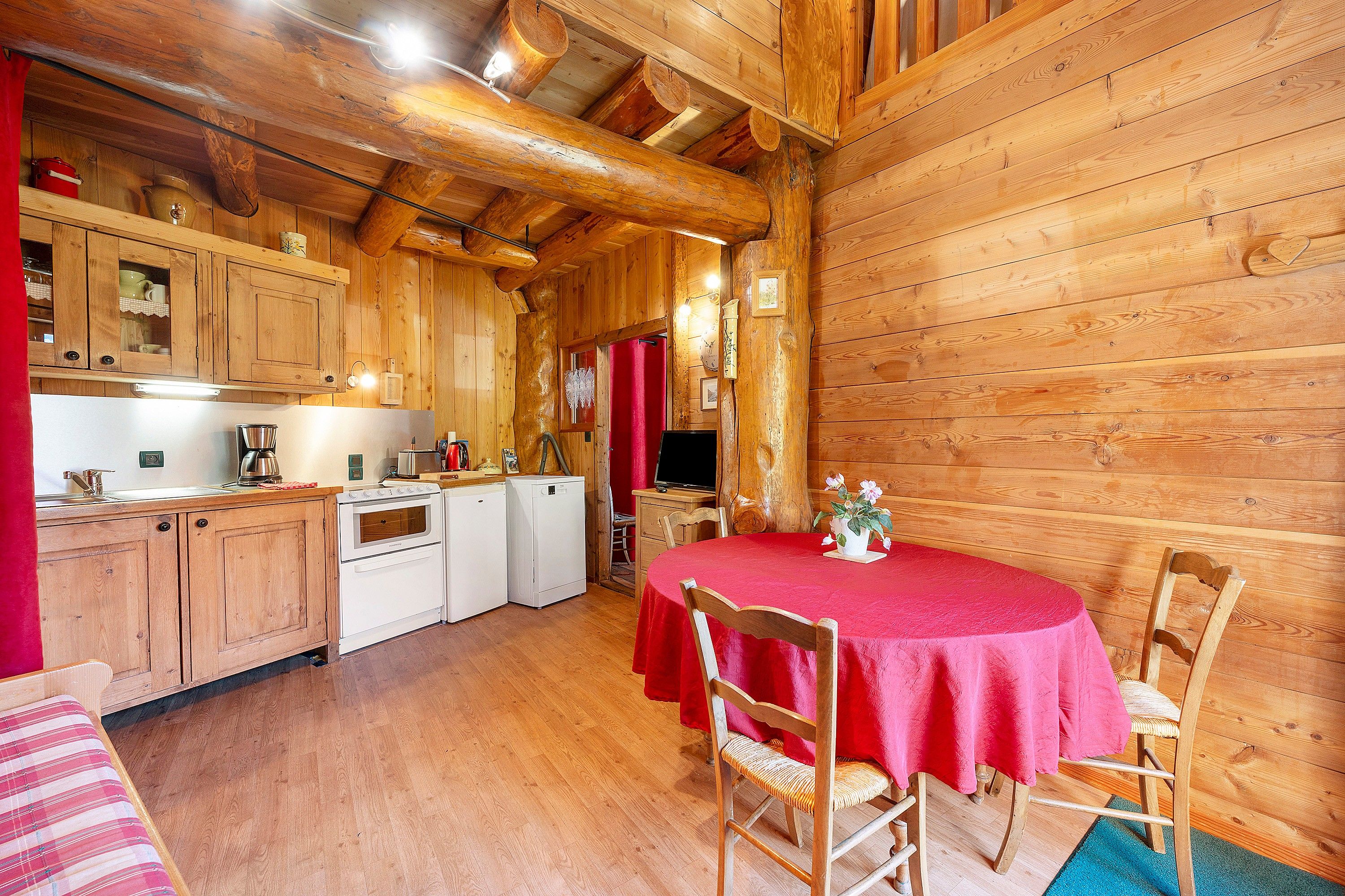 Een gezellig interieur van een chalet met houten wanden, een keuken, een eettafel met stoelen en een rode tafelkleed.