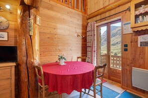 Un interior de madera con una mesa redonda cubierta con un mantel rojo, sillas de madera, un televisor y una ventana con vistas a un paisaje montañoso.