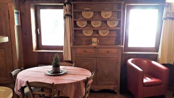 Een houten interieur met een ronde tafel, stoelen, een kast met borden en een rode fauteuil.