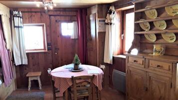 Ein Holzinterieur mit einem runden Tisch, Stühlen, einem kleinen Hocker, einem Schrank mit Geschirr und einem Fenster mit Vorhängen.