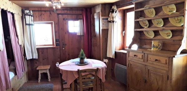 A wooden interior with a round table, chairs, a small stool, a cabinet with dishes, and a window with curtains. - Gîte Les Croës – Chalet Les Abeillos (Bonneval-sur-Arc)
