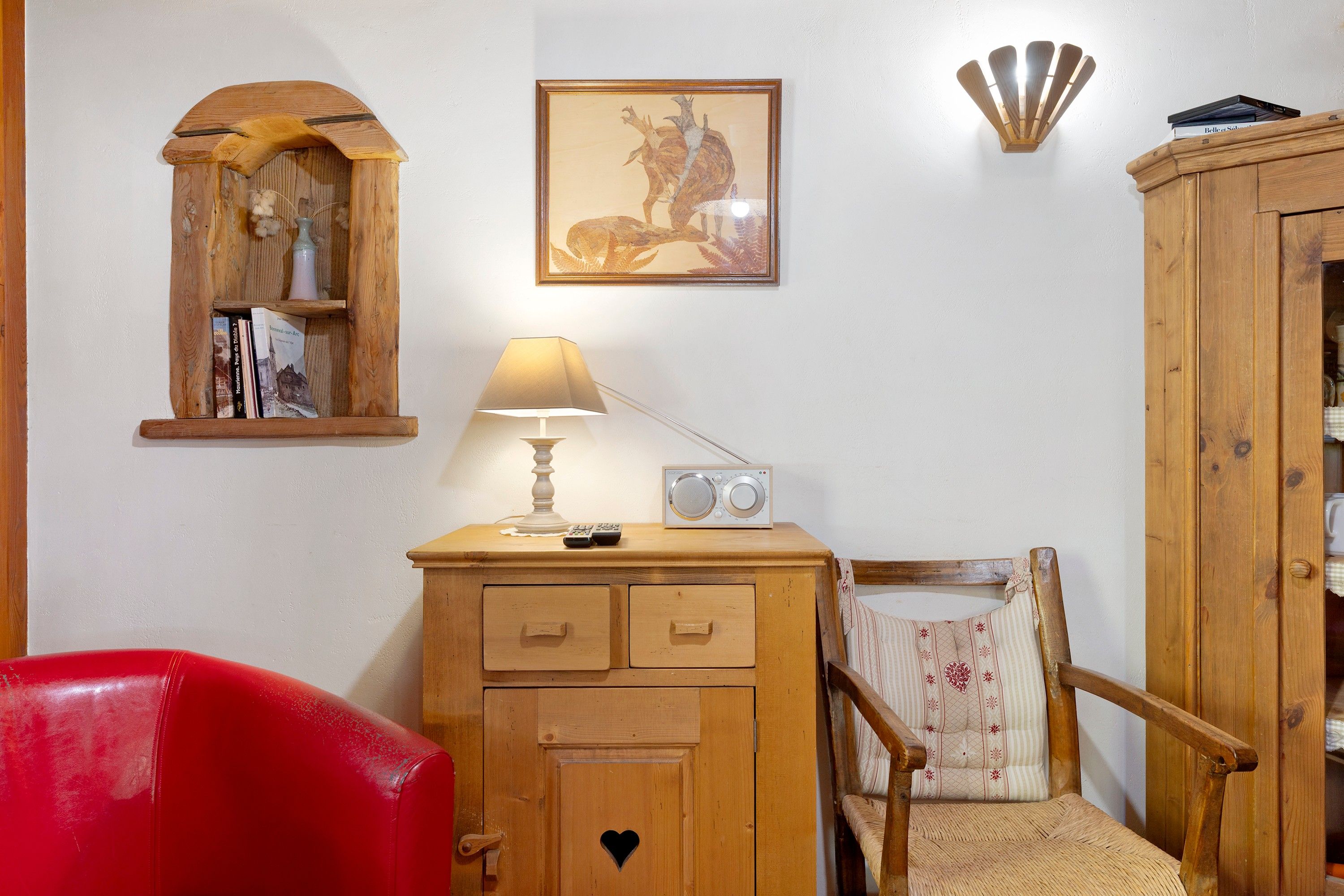 Een kamer met een houten kast, een rode stoel, een lamp, een radio en een ingelijst schilderij aan de muur.