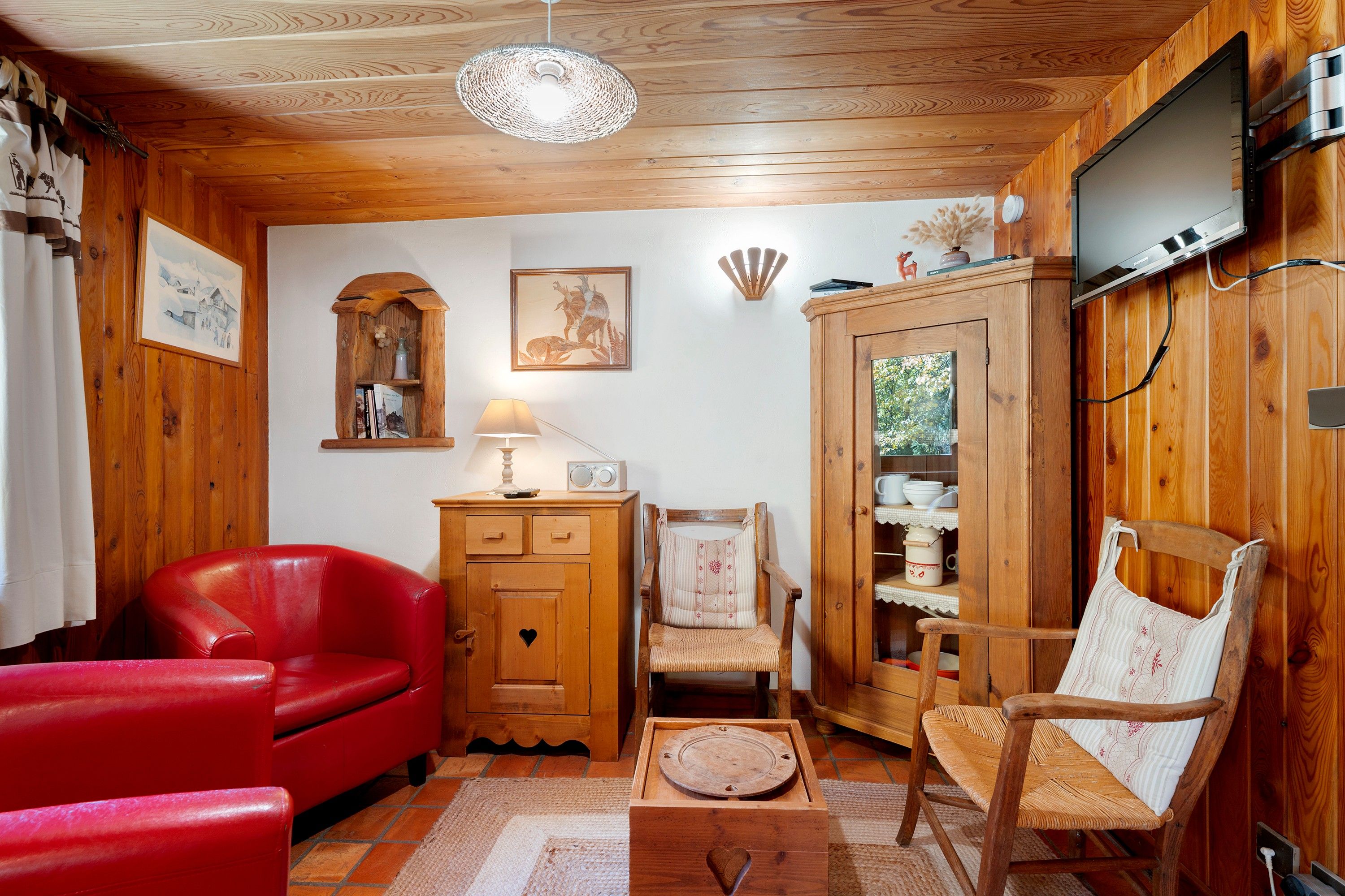A cozy room with wooden walls, a red leather sofa, a wooden cabinet, a wicker chair, and a television mounted on the wall.