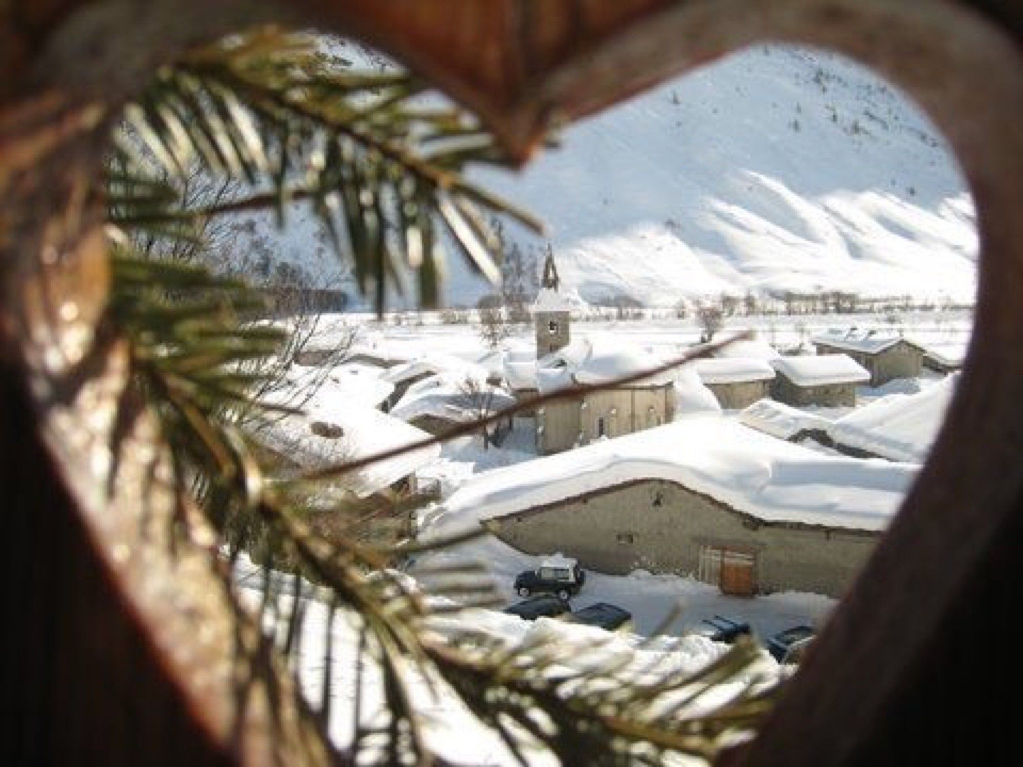 Een besneeuwd dorpsgezicht te zien door een raamkozijn in de vorm van een hart, met dennentakken.