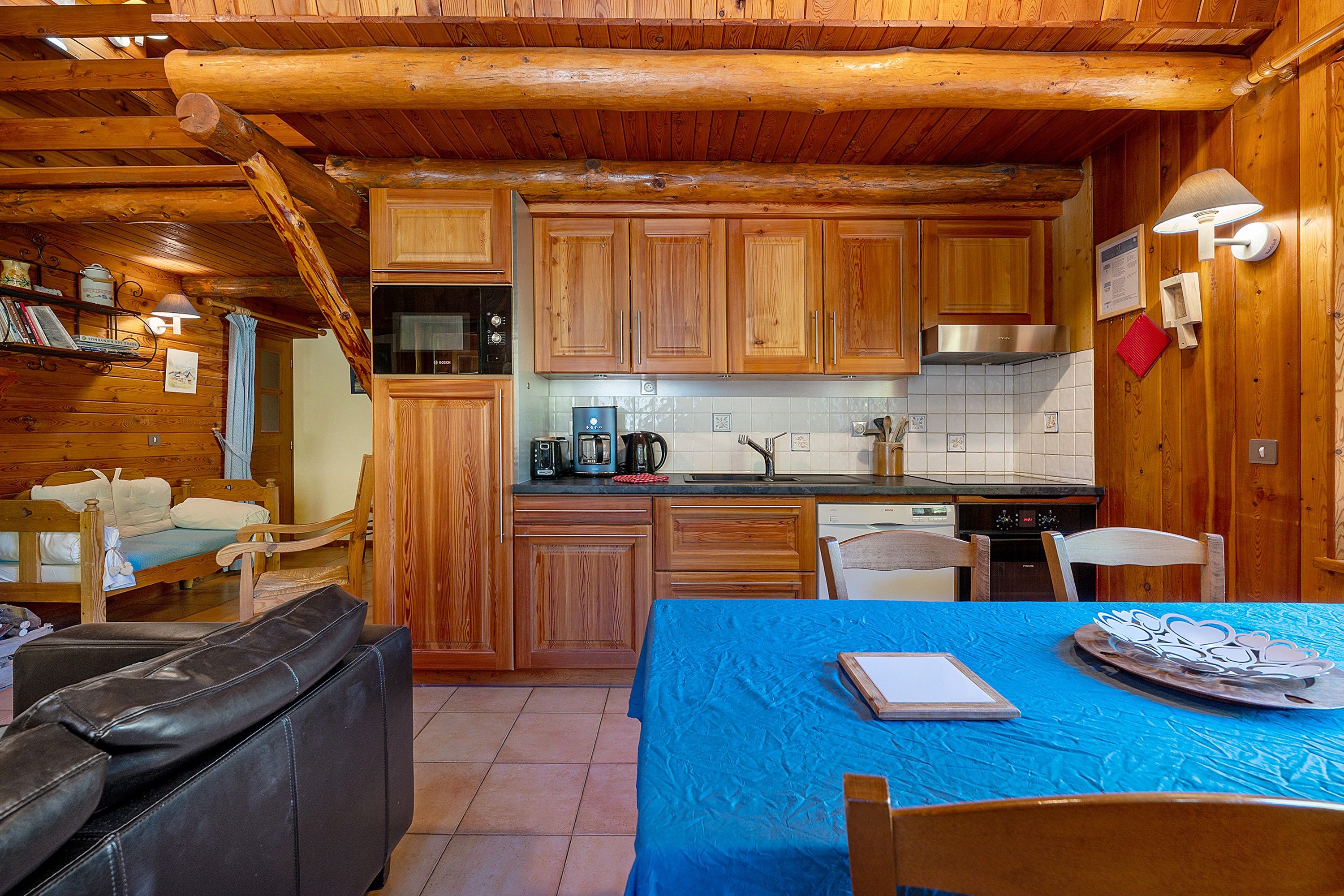 Une cuisine chaleureuse, aménagée dans le style d’un chalet, avec des meubles en bois, une table à manger recouverte d’une nappe bleue et un coin cuisine.