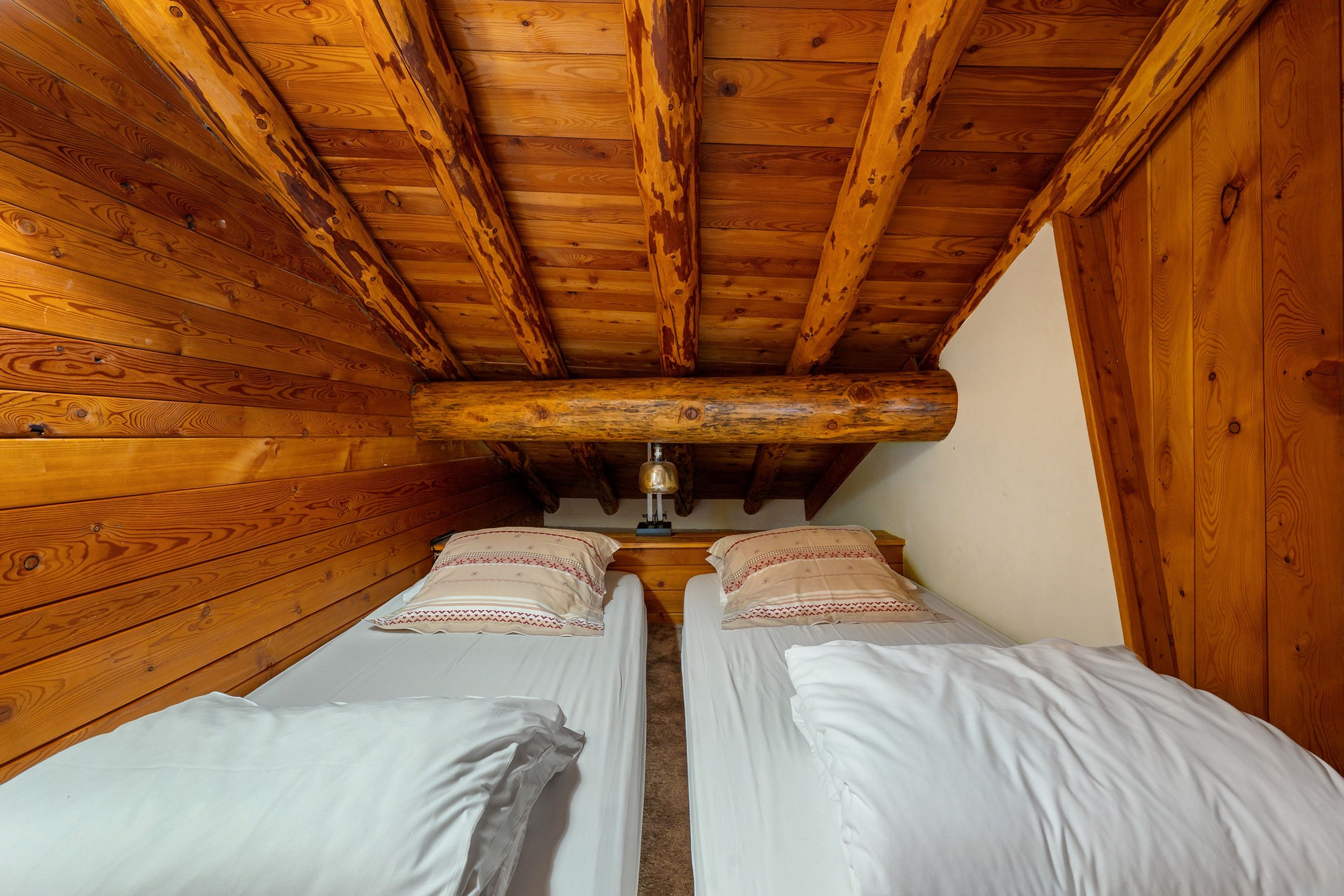 A small, narrow room with wooden walls and a sloped ceiling. There are two beds with white linens and decorative pillows.