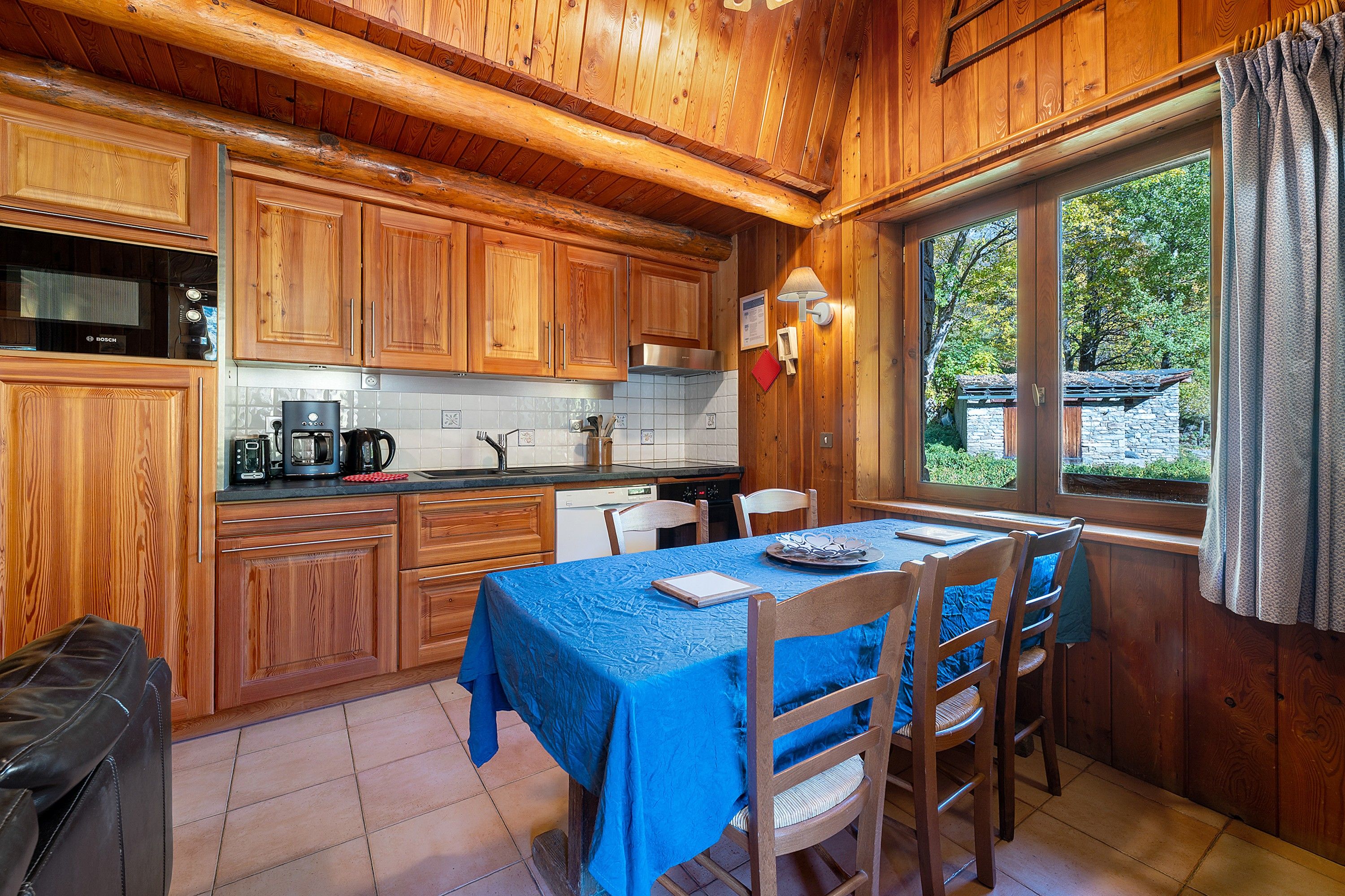 A wooden cabin-style kitchen with a dining area, featuring a table set for four, a Bosch microwave, and a window with curtains.