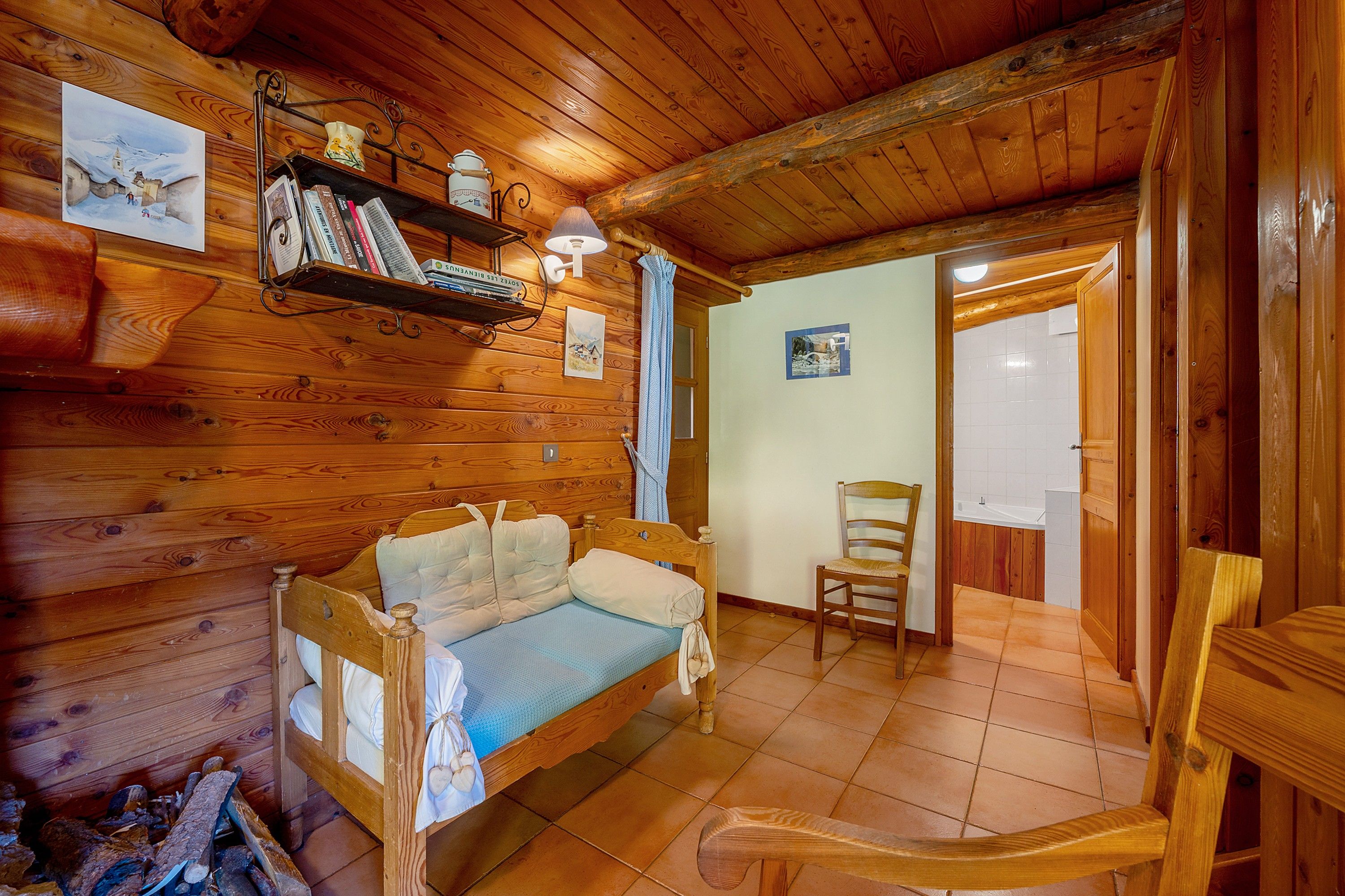 A cozy wooden cabin room with a bed, a chair, a shelf with books, and a painting on the wall.