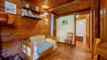 A cozy wooden cabin room with a bed, a chair, a shelf with books, and a painting on the wall.
