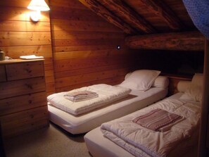 Ett litet, ombonat rum med träväggar och ett sluttande tak. Det finns två enkelsängar med vita sängkläder och en liten byrå med en lampa.