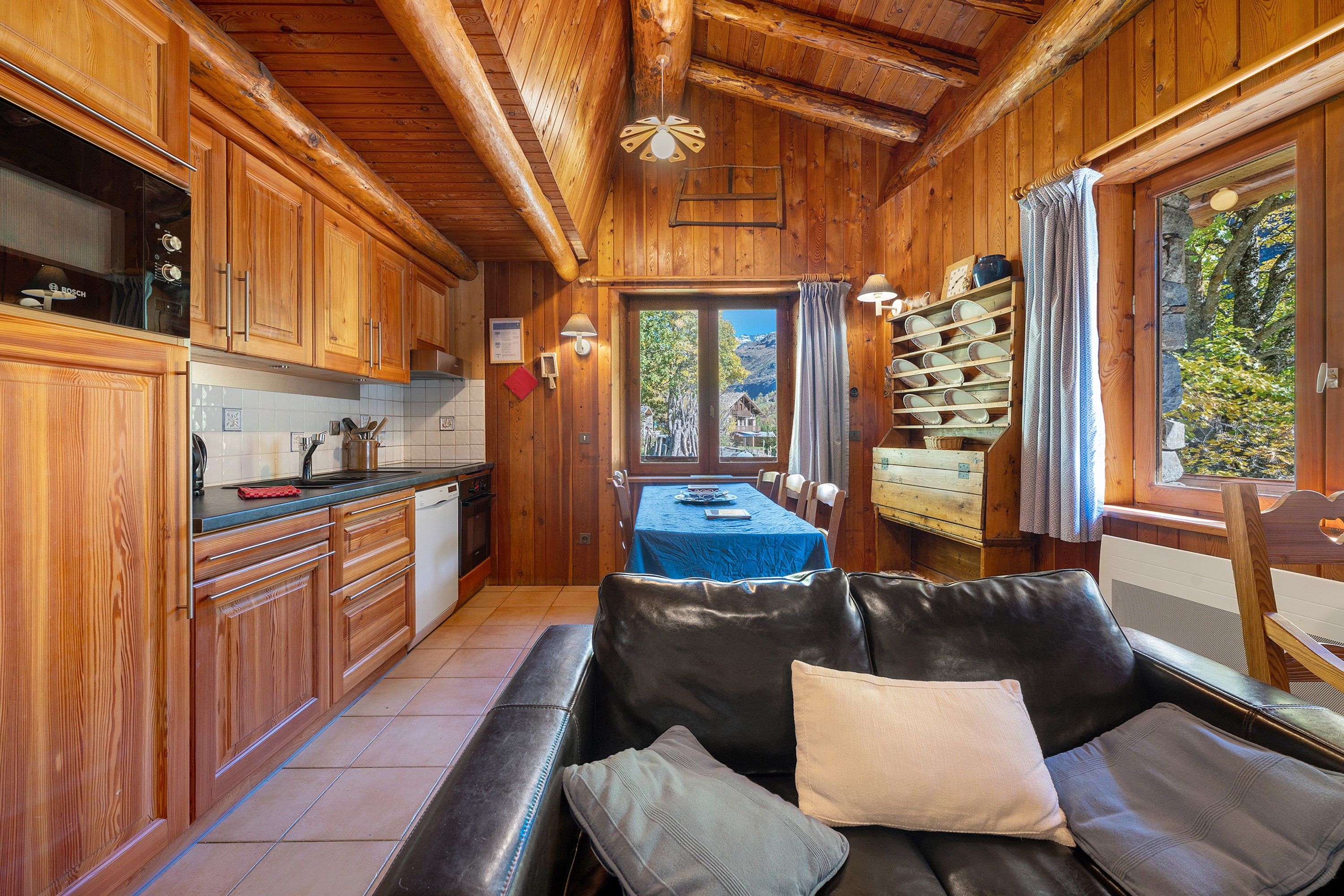 L'intérieur d'un chalet en bois comprend un coin cuisine, un espace repas avec une table et des chaises, ainsi qu'un canapé en cuir agrémenté de coussins.