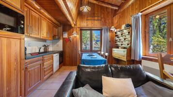 L'intérieur d'un chalet en bois comprend un coin cuisine, un espace repas avec une table et des chaises, ainsi qu'un canapé en cuir agrémenté de coussins.