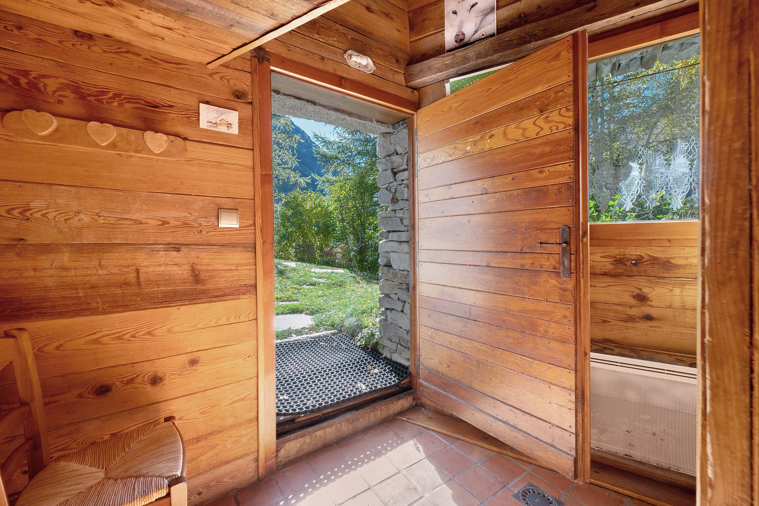 L’intérieur d’un chalet en bois, avec une entrée en pierre, un petit coin salon et une vue sur un jardin et des montagnes.