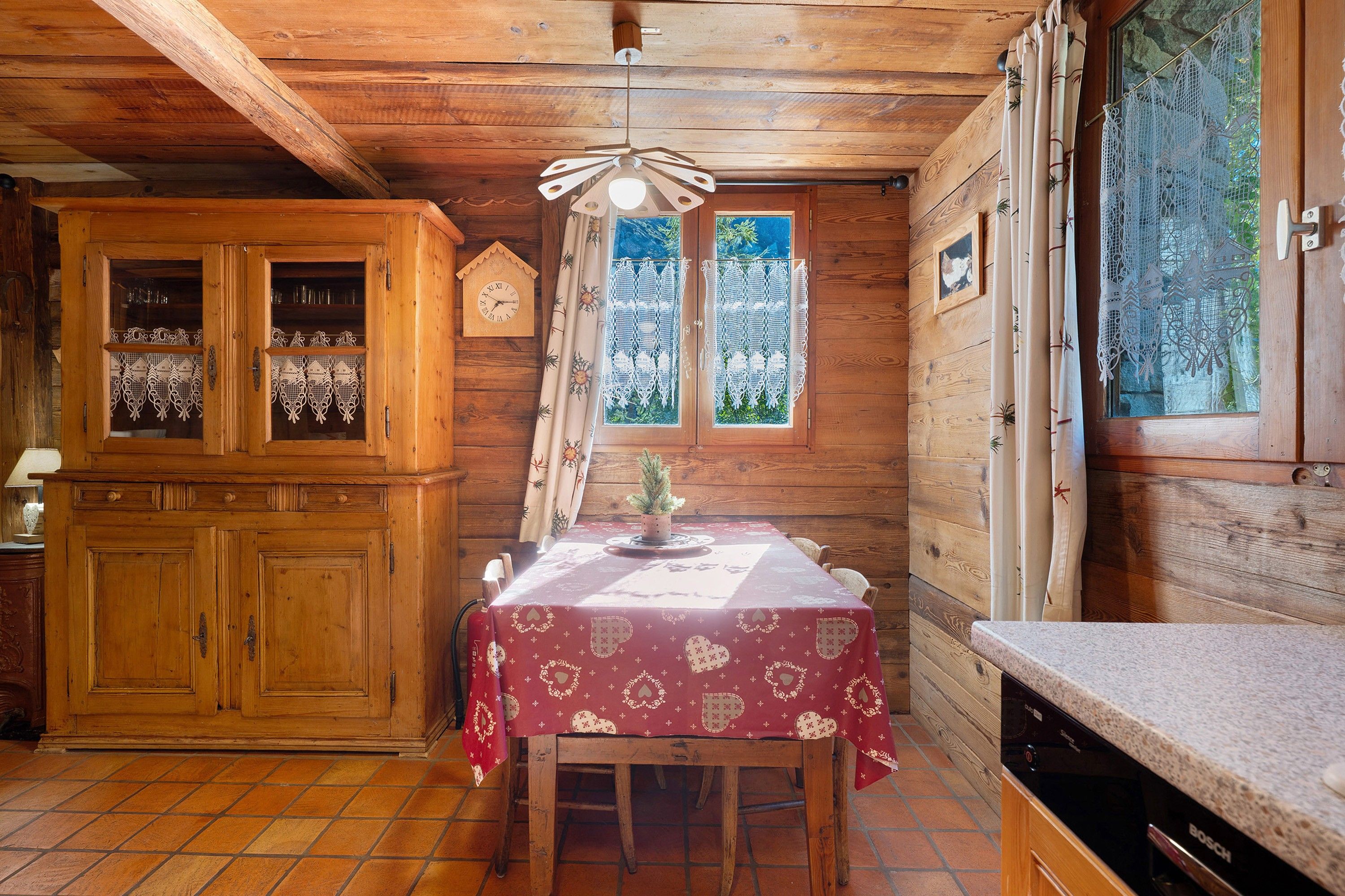 Ein Interieur einer Holzhütte mit Esstisch, Holzschränken und einem Fenster mit Spitzenvorhängen.