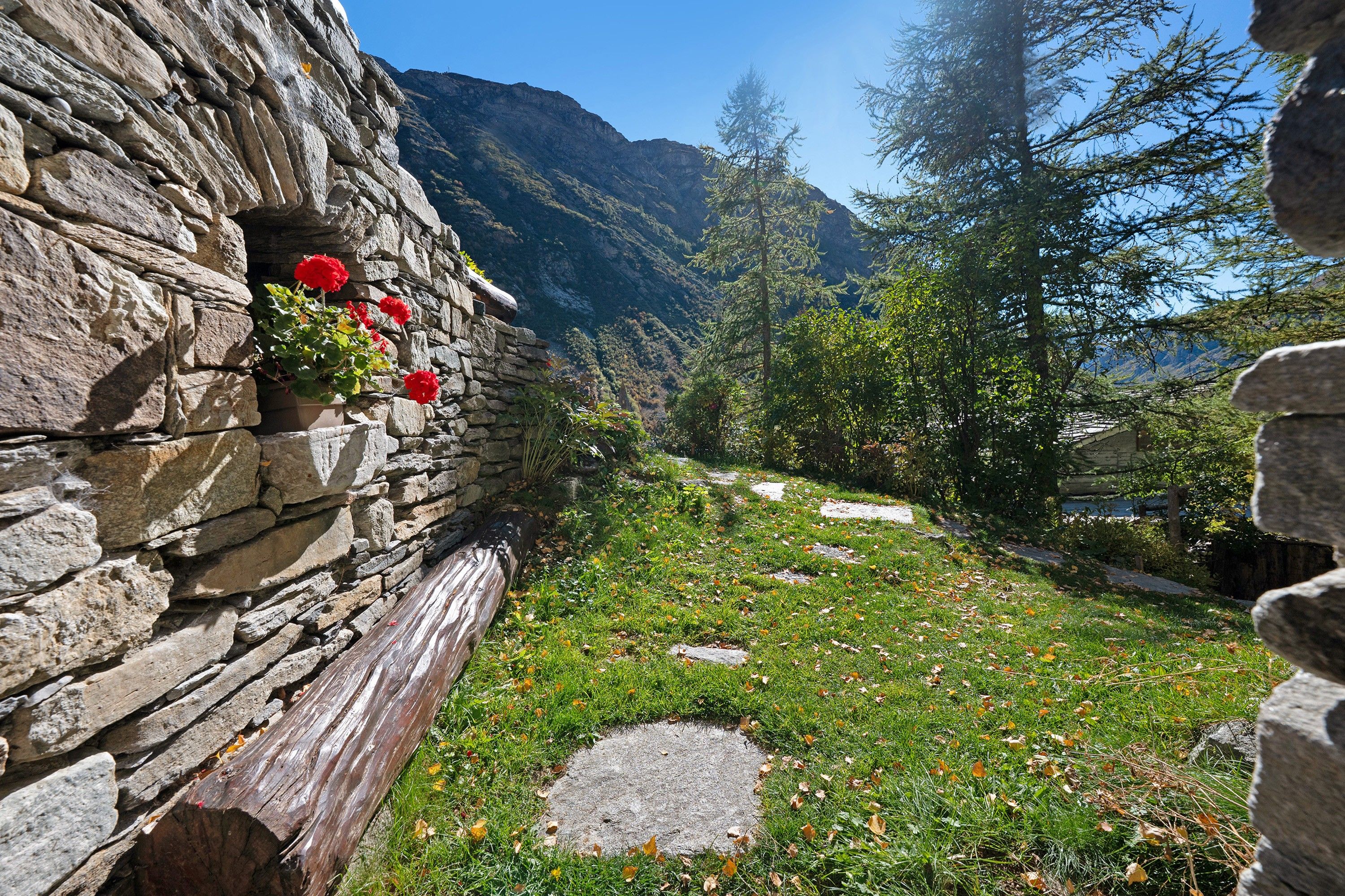 Un mur de pierre agrémenté d’une jardinière et un sentier qui traverse une zone herbeuse, avec des arbres et des montagnes en arrière-plan.