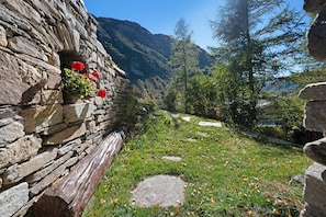 A stone wall with a flower box and a pathway leading through a grassy area with trees and mountains in the background. - Gîte l'Erablo – Chalet Les Abeillos in Bonneval-sur-Arc, Vanoise National Park (Bonneval-sur-Arc)
