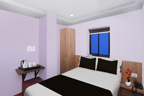A hotel room with a bed, a desk with a kettle and cups, a wardrobe, a window with a blue view, and a phone on the nightstand.