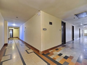 A long, narrow hallway with a patterned floor, doors on one side, and a plain wall on the other.
