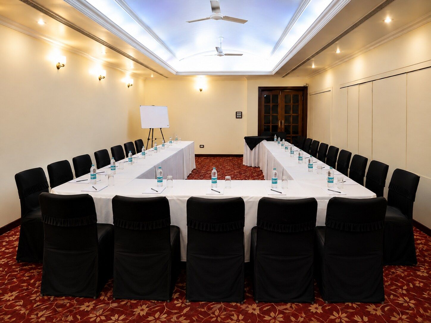 Un salón de conferencias con una larga mesa, sillas negras, botellas de agua y una pizarra.