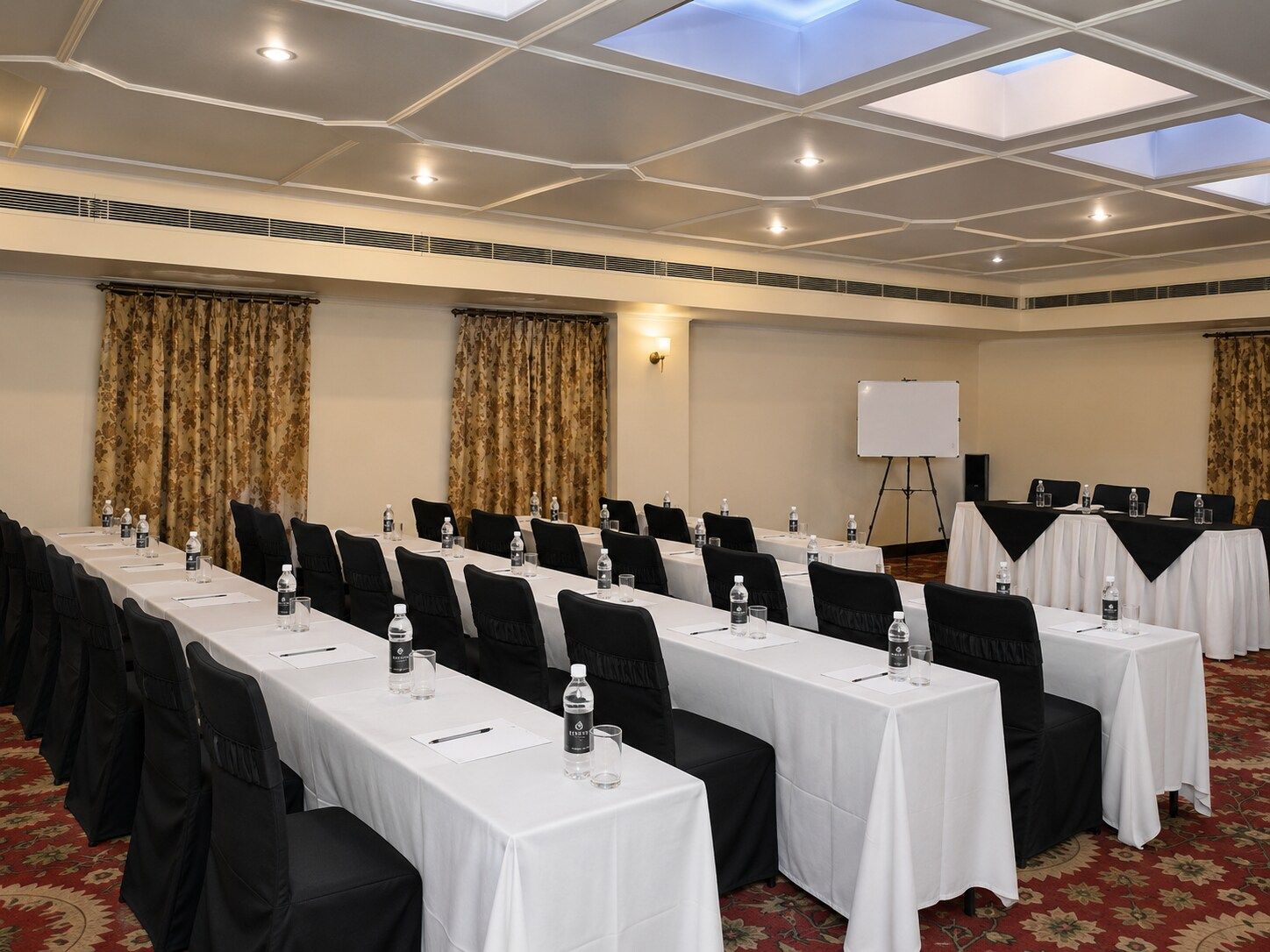 Un salón de conferencias con mesas largas, sillas negras, manteles blancos, botellas de agua y una pizarra.
