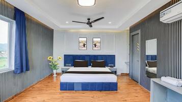 Et hotelværelse med en stor seng, ventilator i loftet, et vindue med gardiner, en vægmonteret aircondition og et lille bord med blomster i en vase.