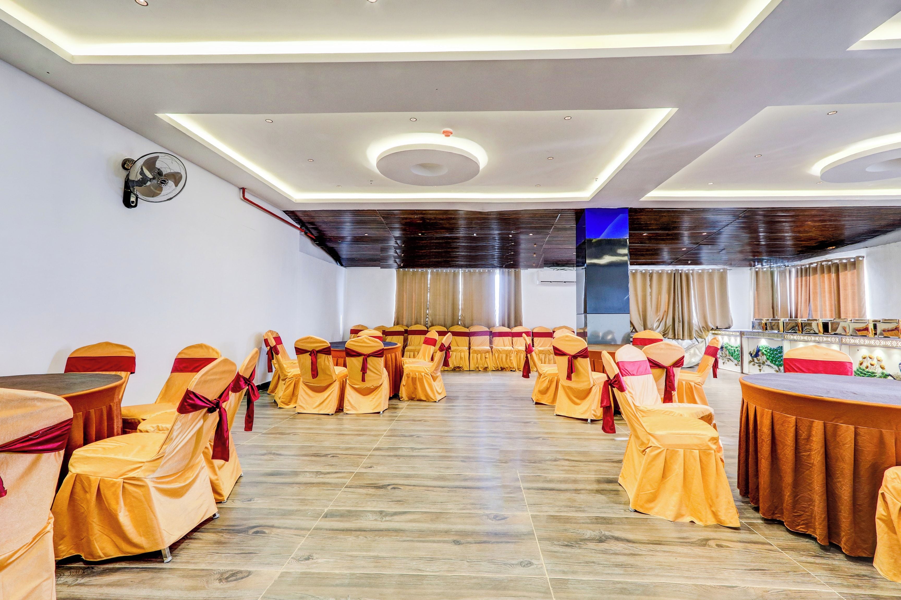 Uma sala de conferências com fileiras de mesas e cadeiras, um ventilador de teto e uma grande janela com cortinas.