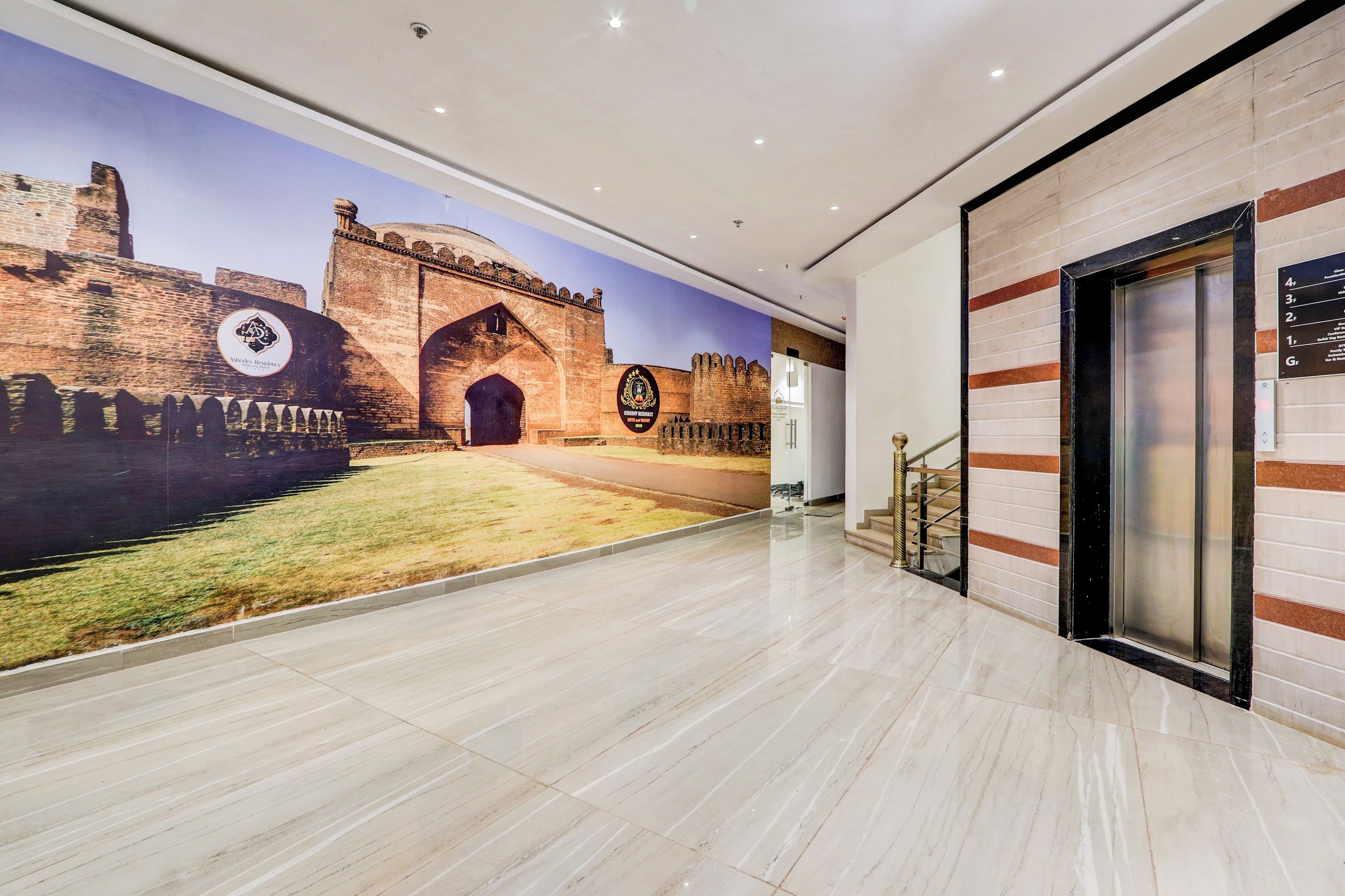 Een hotel lobby met een groot muurdecoratie van een historisch gebouw, een moderne lift en een bord dat de locatie van Ashodey Residency aangeeft.