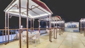 Un espacio de estar al aire libre techado con mesas y sillas, barandilla y vista nocturna de la ciudad.
