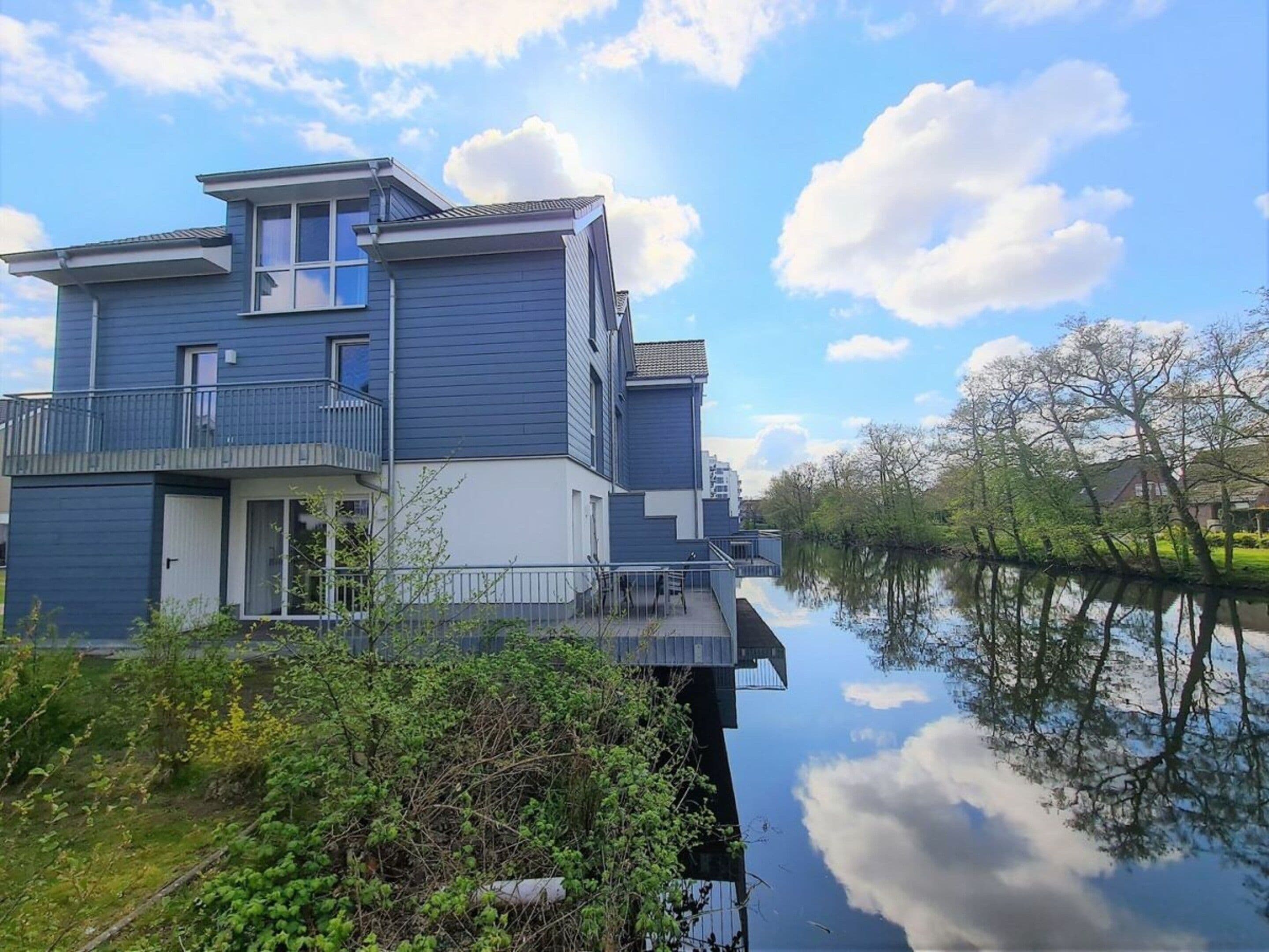 A modern two-story house with a balcony, situated next to a calm body of water, reflecting the sky and surrounding trees.