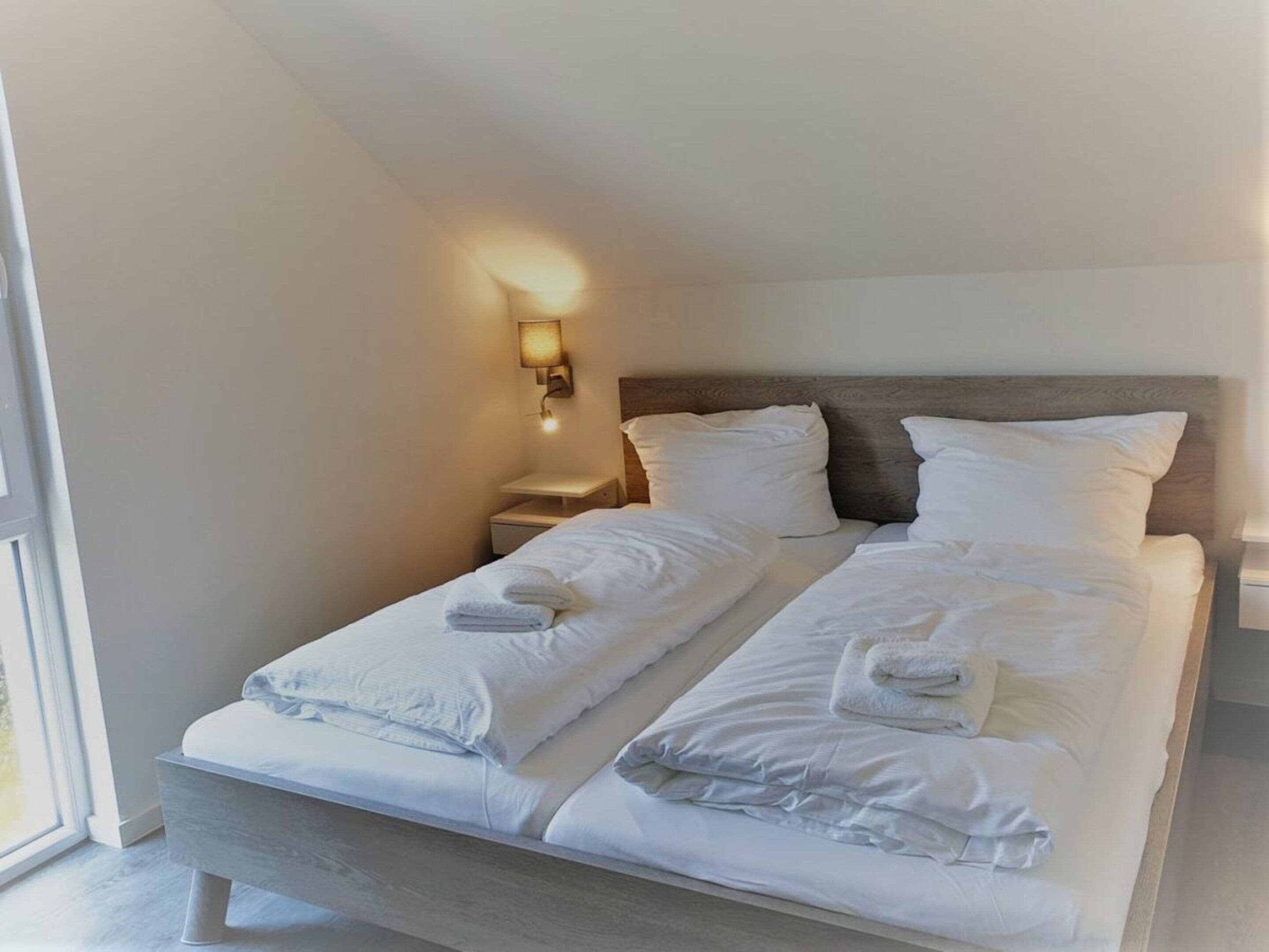 A double bed with white linens and towels, a bedside table, a nightstand, and a wall-mounted lamp.
