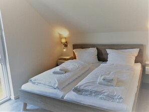 A double bed with white linens and towels, a bedside table, a nightstand, and a wall-mounted lamp. - House No. 33, Holiday Home Deichgraf (Wangerland)