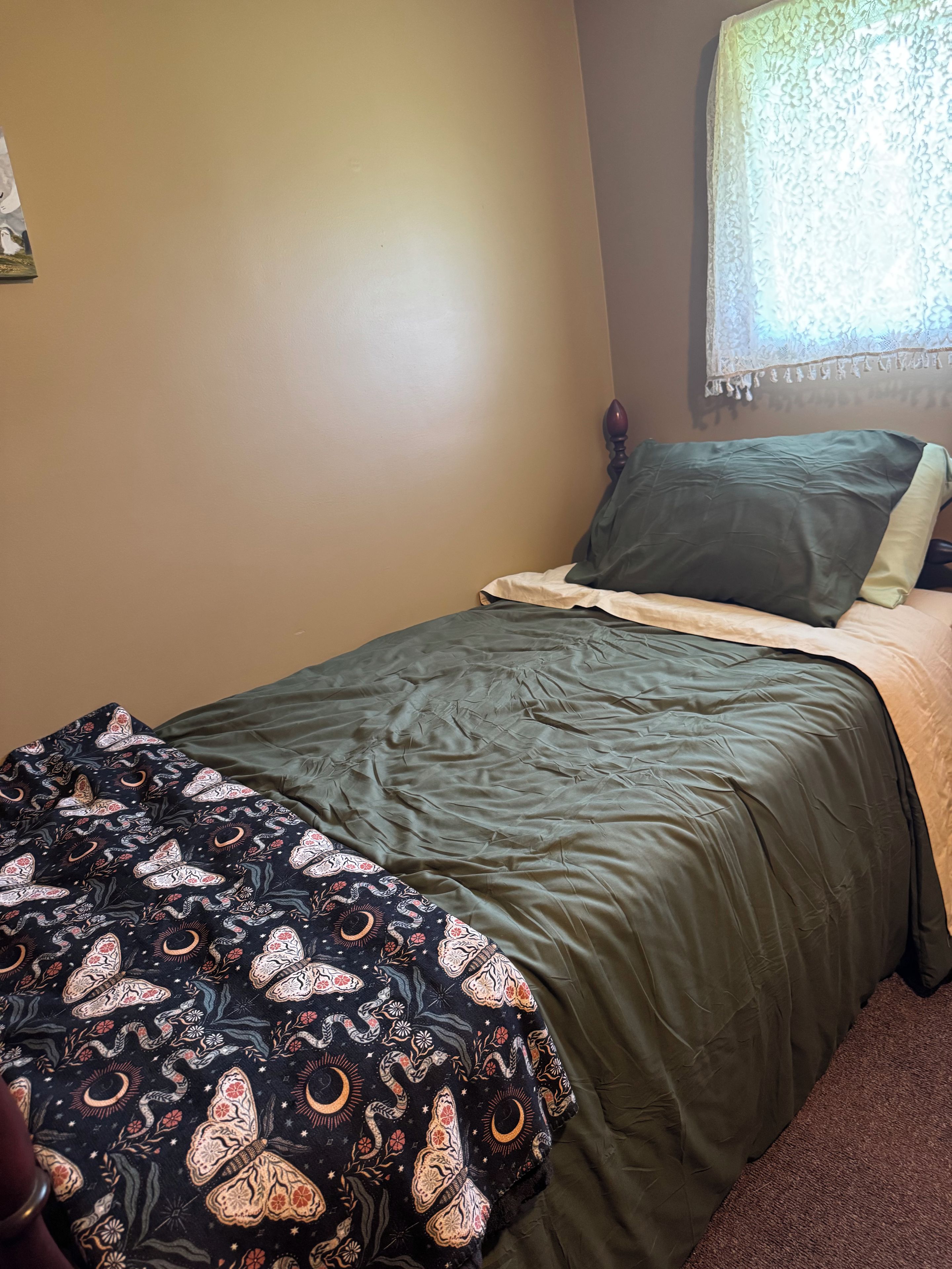 A bedroom with a bed, a window with lace curtains, and a wall with a floral pattern.
