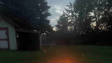 Un foyer extérieur circulaire, avec des flammes, entouré de dalles de pierre, situé dans une cour verdoyante avec des arbres et un bâtiment en arrière-plan.
