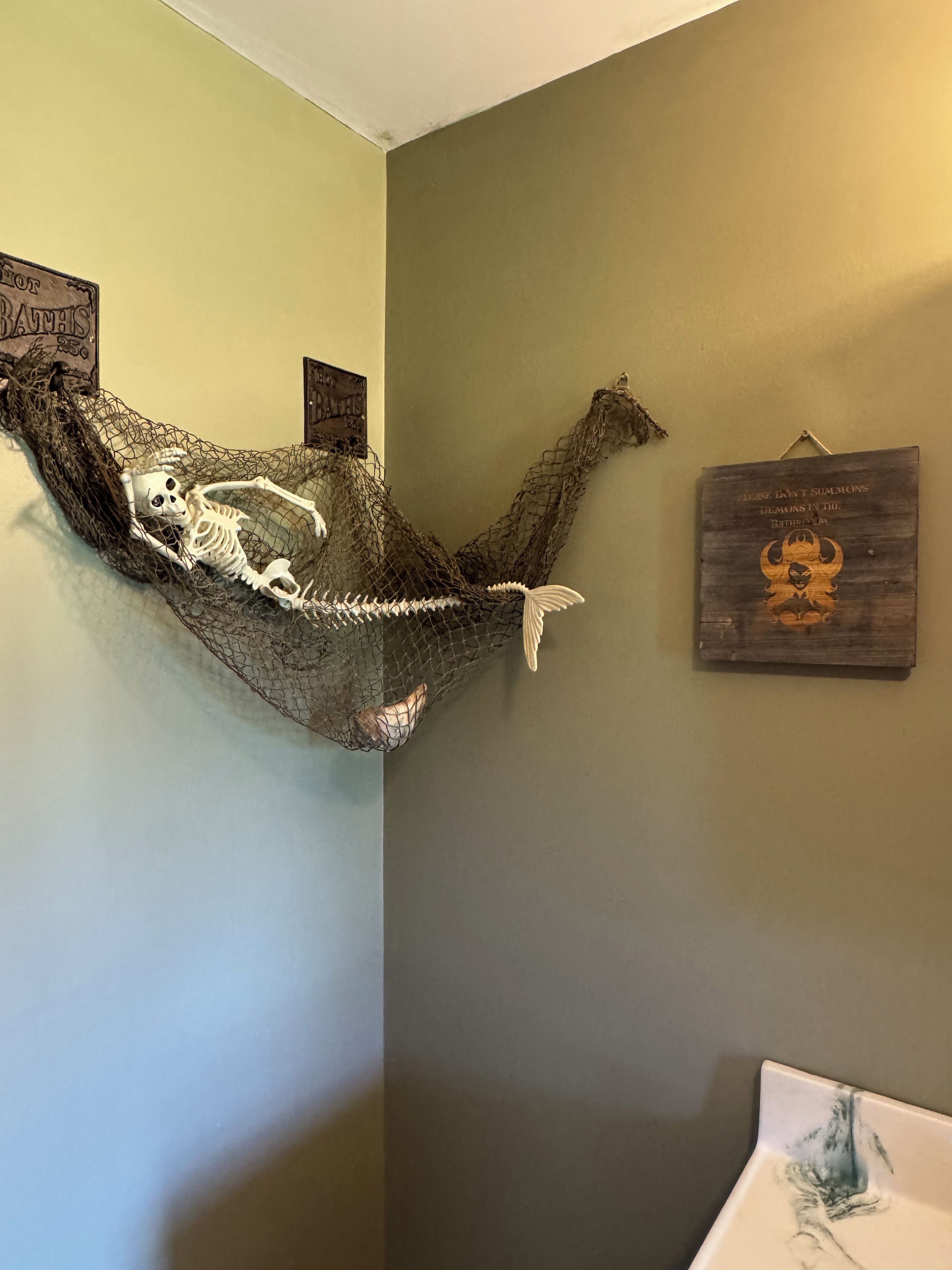 A room with a skeleton in a hammock, a wooden sign with a skull, and a wall-mounted picture.