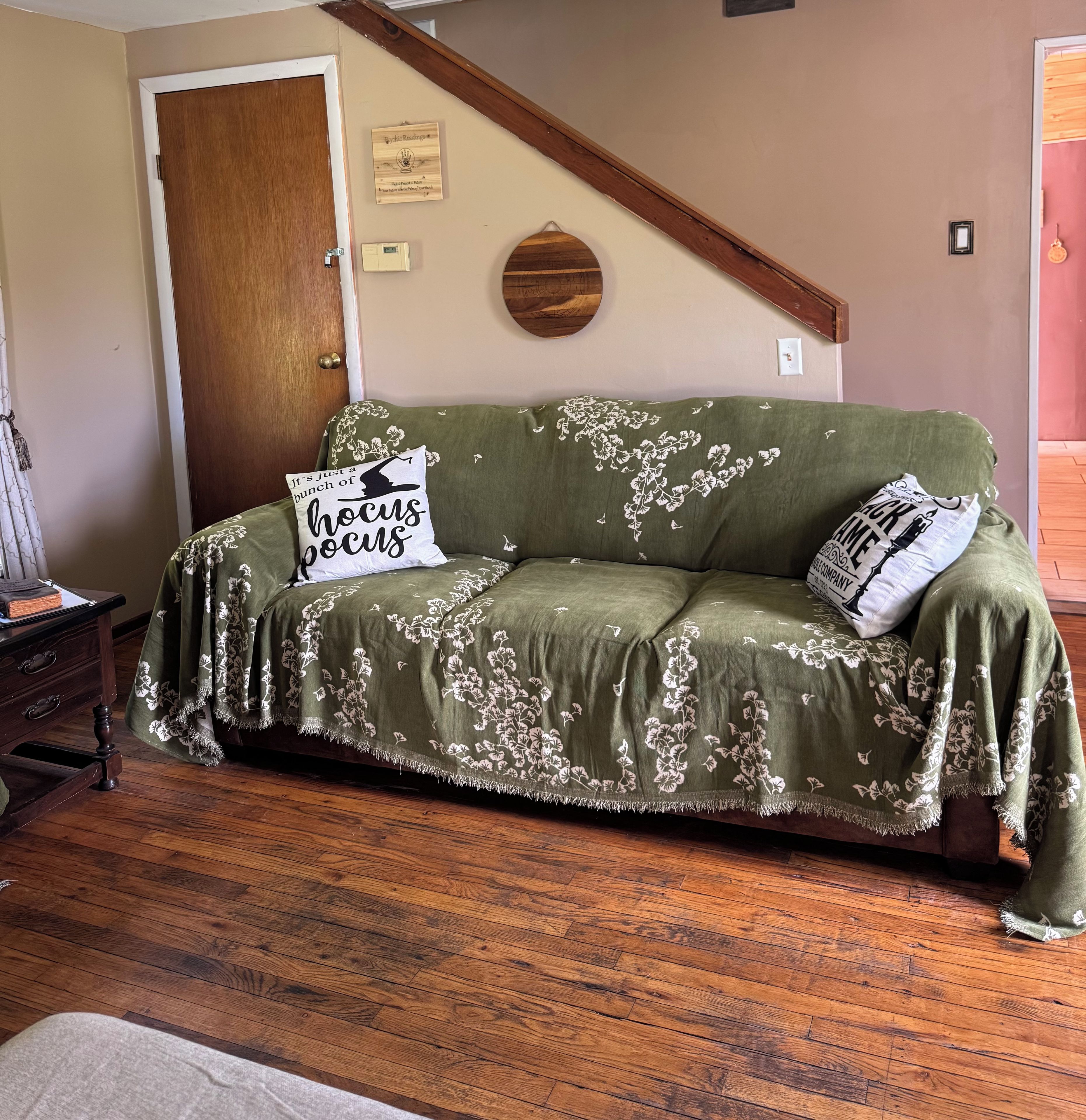 A green sofa with floral patterns and two pillows, one with 'hocus pocus' text, in a room with wooden flooring and a wooden door.