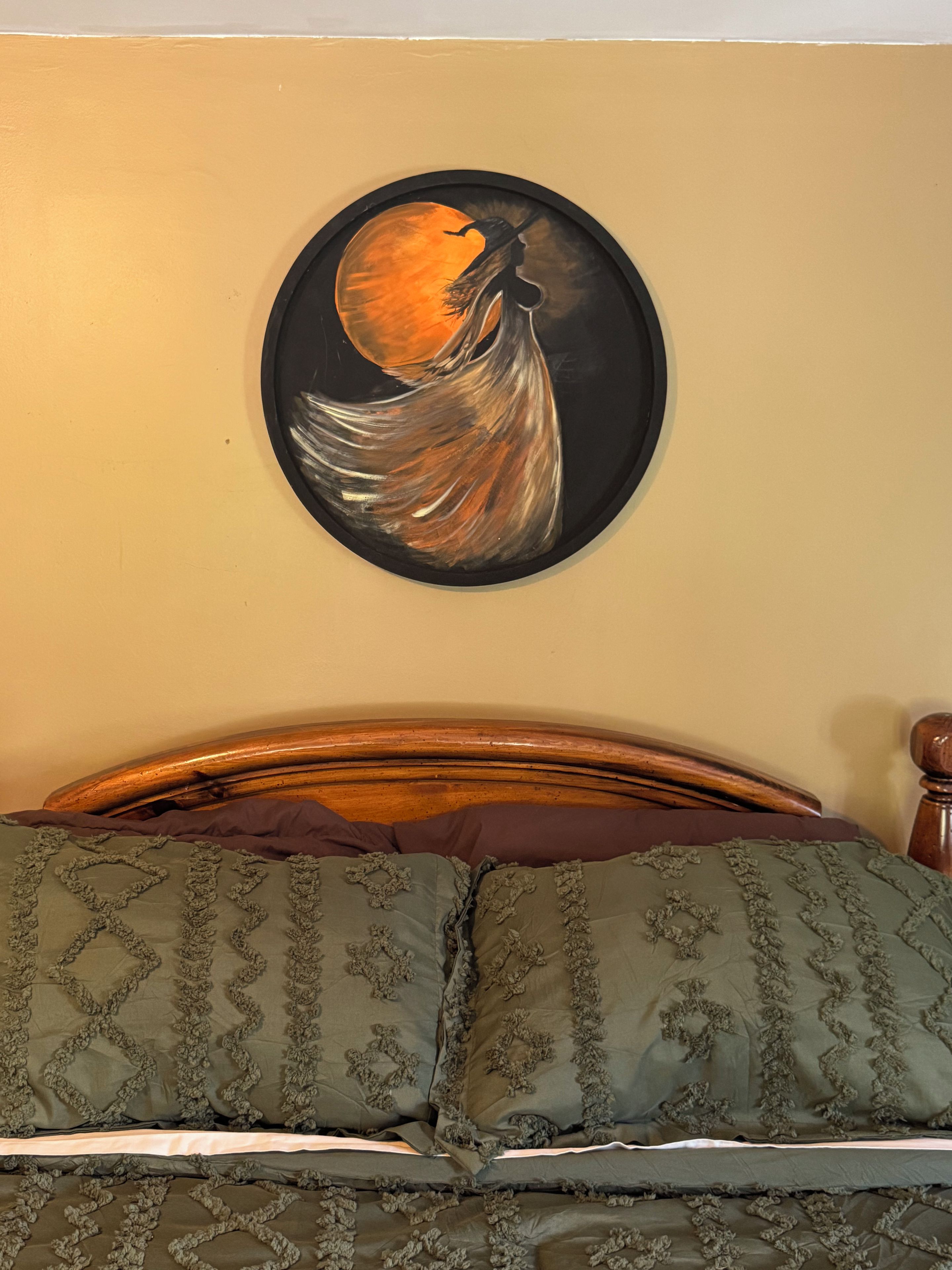 Un letto con una testata in legno, un cuscino a fantasia verde e un quadro appeso al muro raffigurante una figura con veste fluente su sfondo circolare.