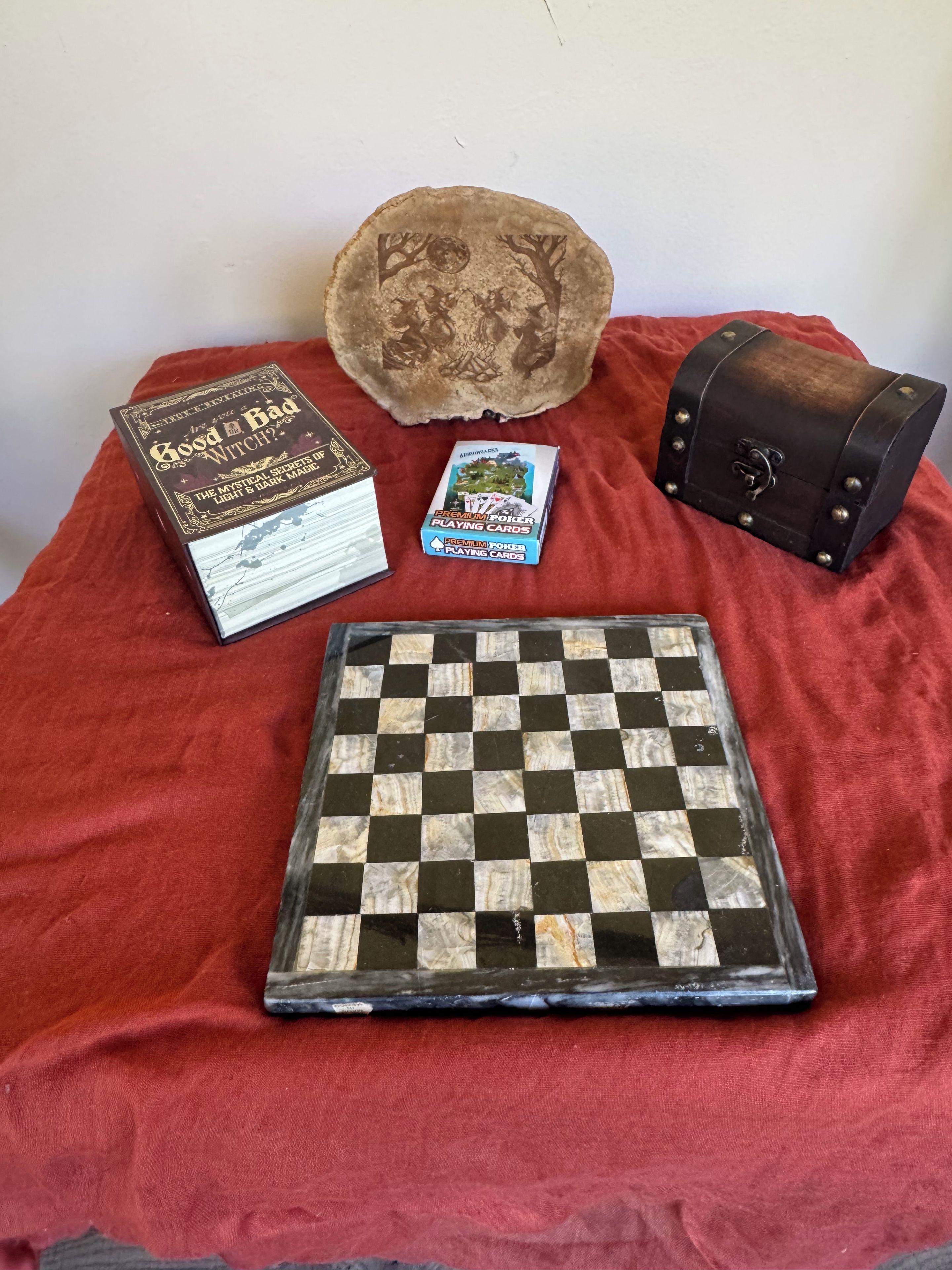 Un jeu d’échecs, un livre intitulé « La Bonne Sorcière », une boîte de cartes à jouer de qualité supérieure et une petite pochette en cuir sur une surface en tissu rouge.