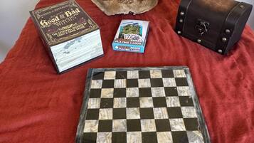 Un jeu d’échecs, un livre intitulé « La Bonne Sorcière », une boîte de cartes à jouer de qualité supérieure et une petite pochette en cuir sur une surface en tissu rouge.