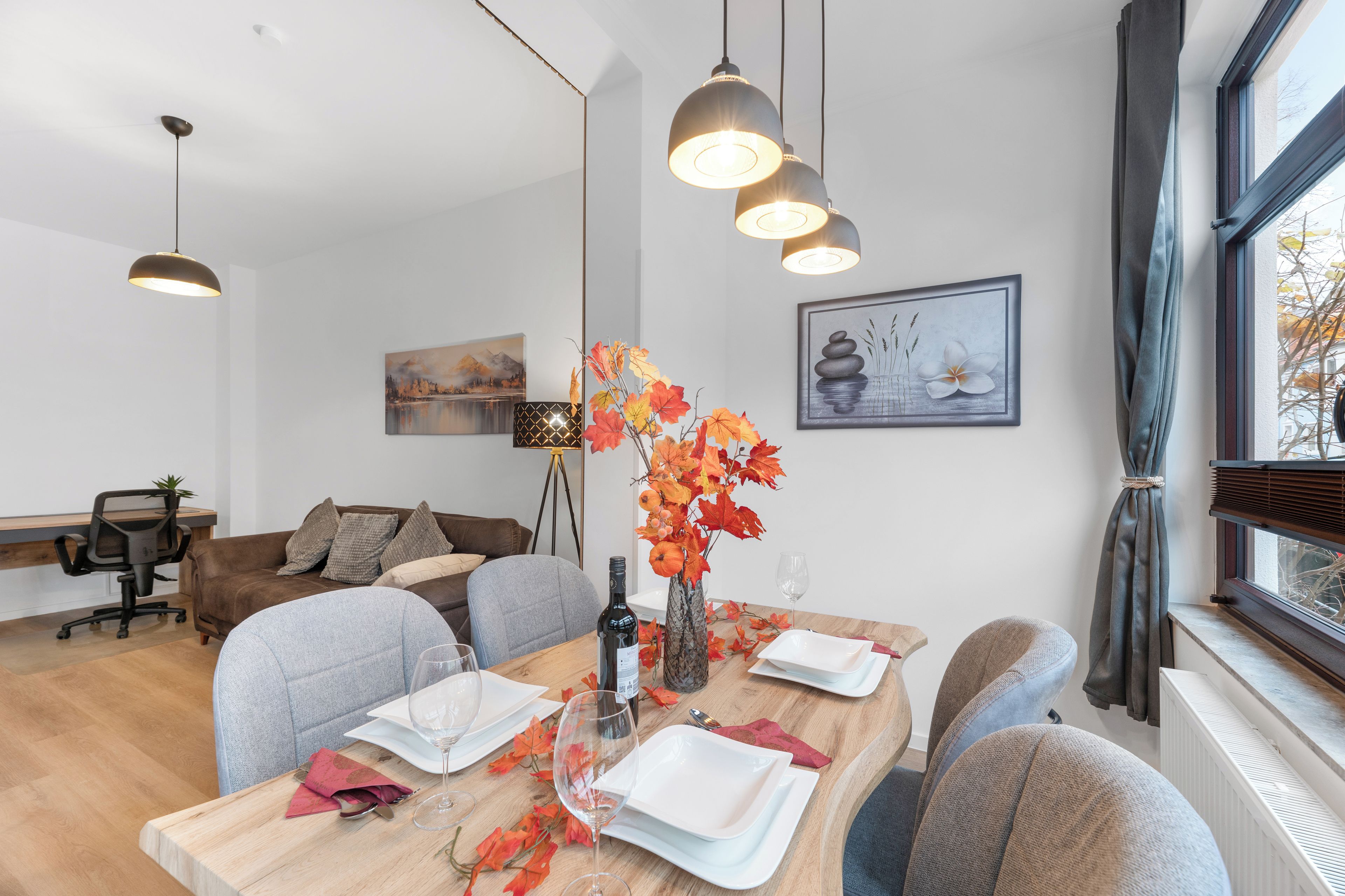 A dining area with a wooden table set for four, a vase with autumn leaves, and a painting on the wall.