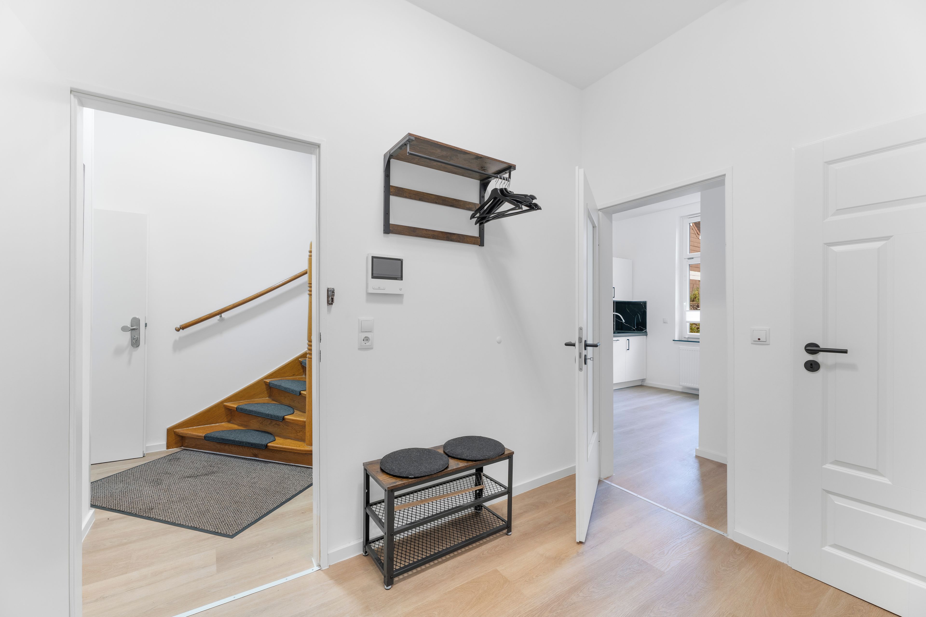 Ein Flur mit einer Holztreppe, einem Kleiderständer, einem kleinen Tisch mit Hocker und einer Tür zu einem anderen Raum.