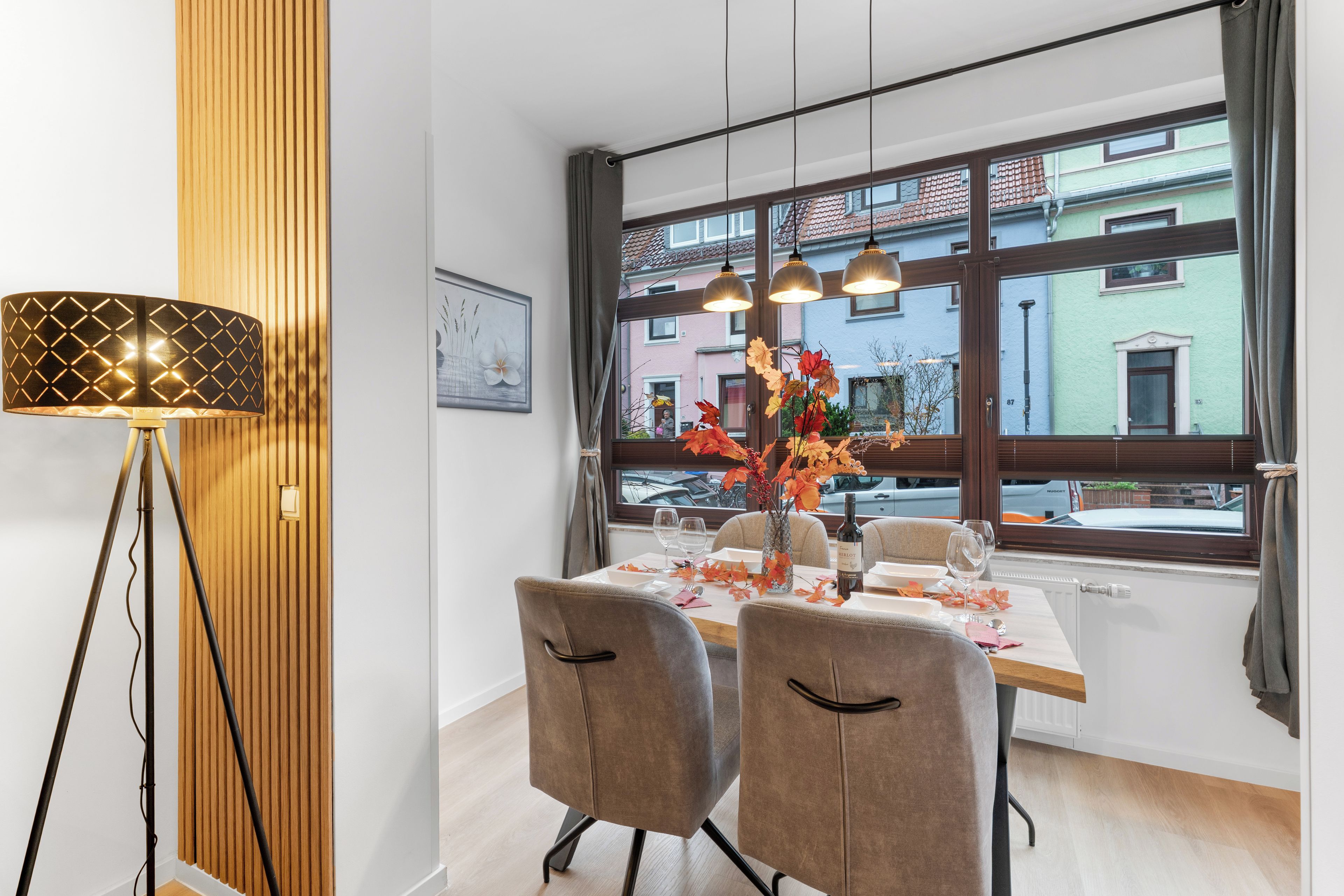A dining area with a table set for two, a vase with autumn leaves, and a view of buildings through large windows.