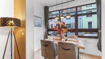 A dining area with a table set for two, a vase with autumn leaves, and a view of buildings through large windows.