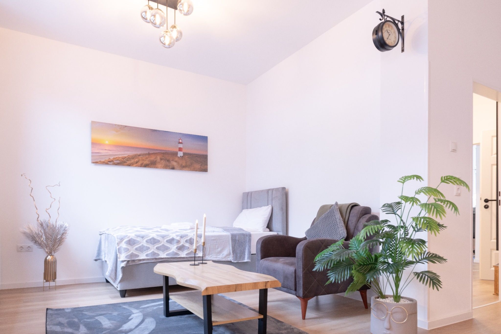 Ein modernes Schlafzimmer mit Bett, Nachttisch, Lampe, Stuhl und einem Gemälde, das einen Sonnenuntergang über einem Strand zeigt.