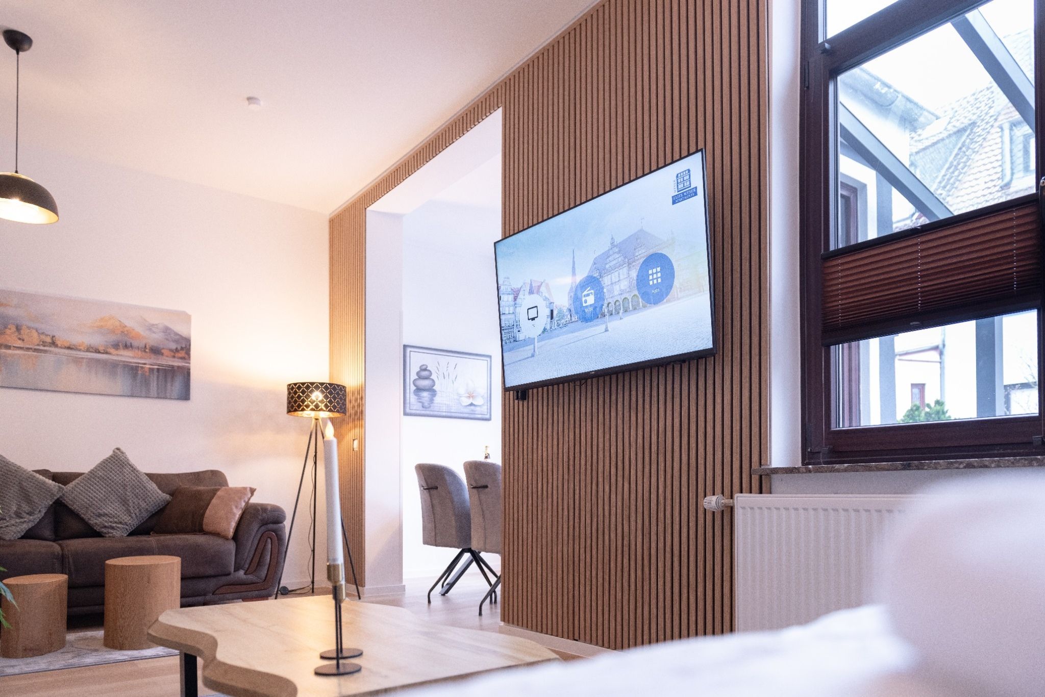 A modern living room with a flat-screen TV mounted on a wooden wall, a sofa, a floor lamp, and a table with chairs.