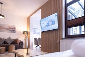 A modern living room with a flat-screen TV mounted on a wooden wall, a sofa, a floor lamp, and a table with chairs.
