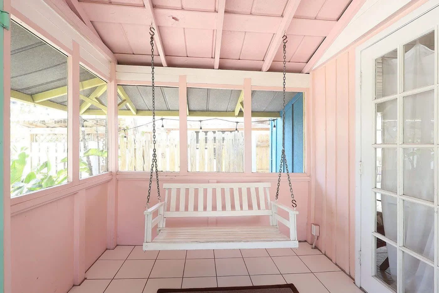 A pink-walled porch with a white swing bench, a tiled floor, and a view of a wooden fence.